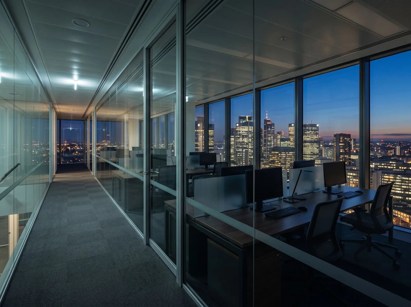 Andar de escritório corporativo ao anoitecer, divisórias de vidro e cidade ao fundo.